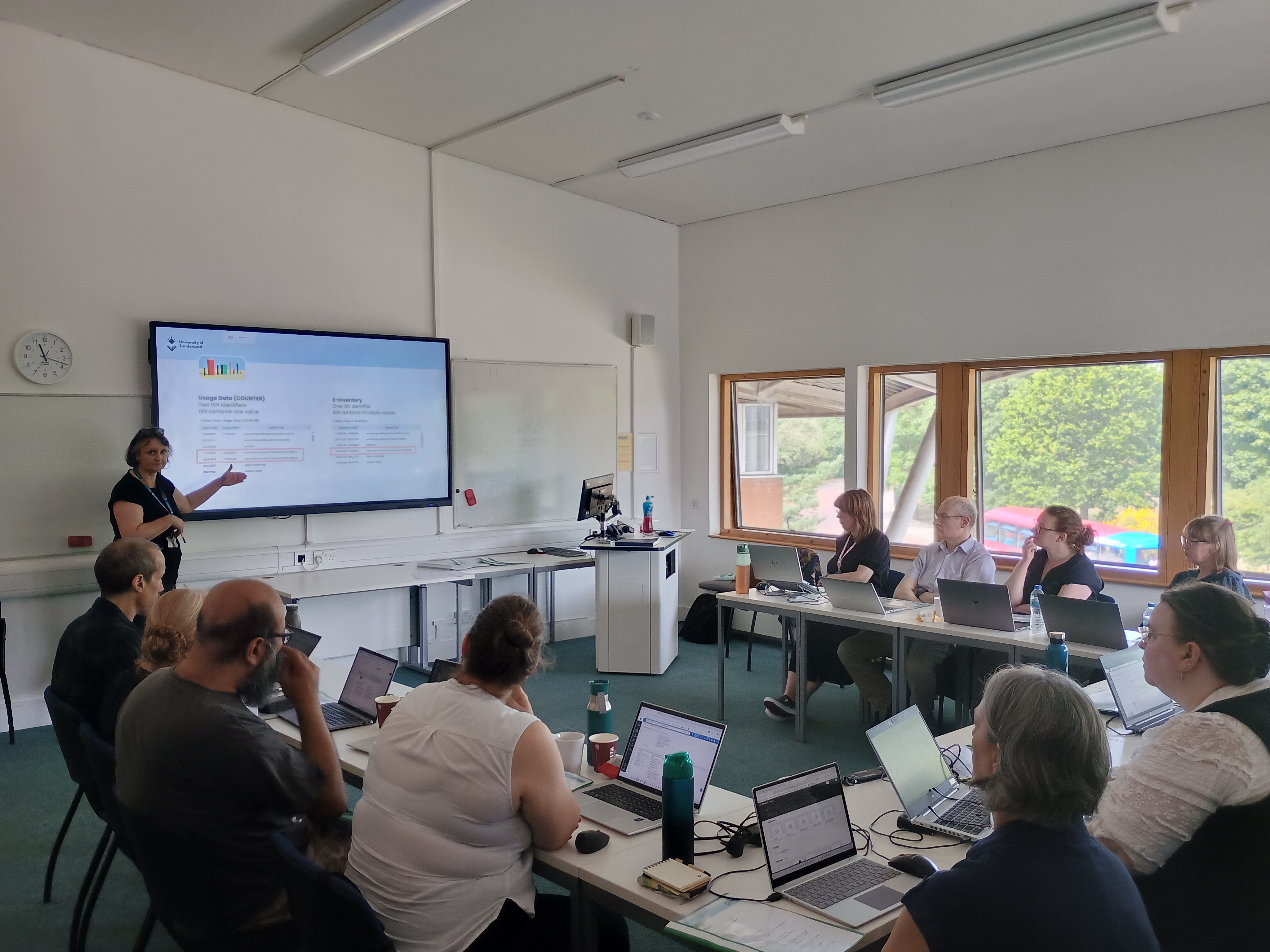 A classroom of workshop participants seated in a horseshoe shape in front of a screen with an instructor pointing